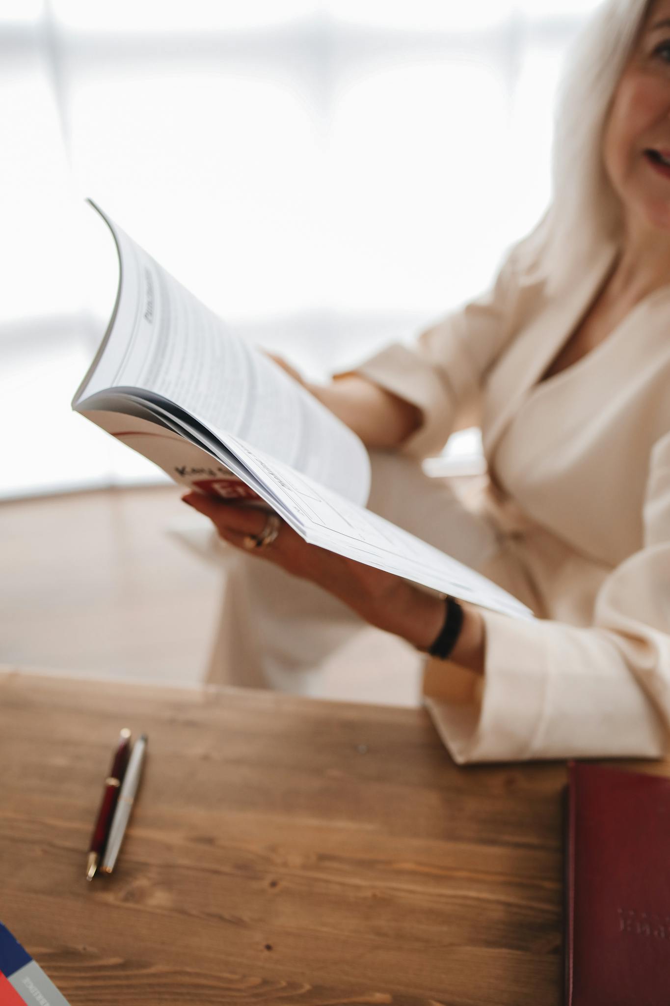 A woman in light clothing reads a book at a wooden desk in a well-lit room, exemplifying learning.