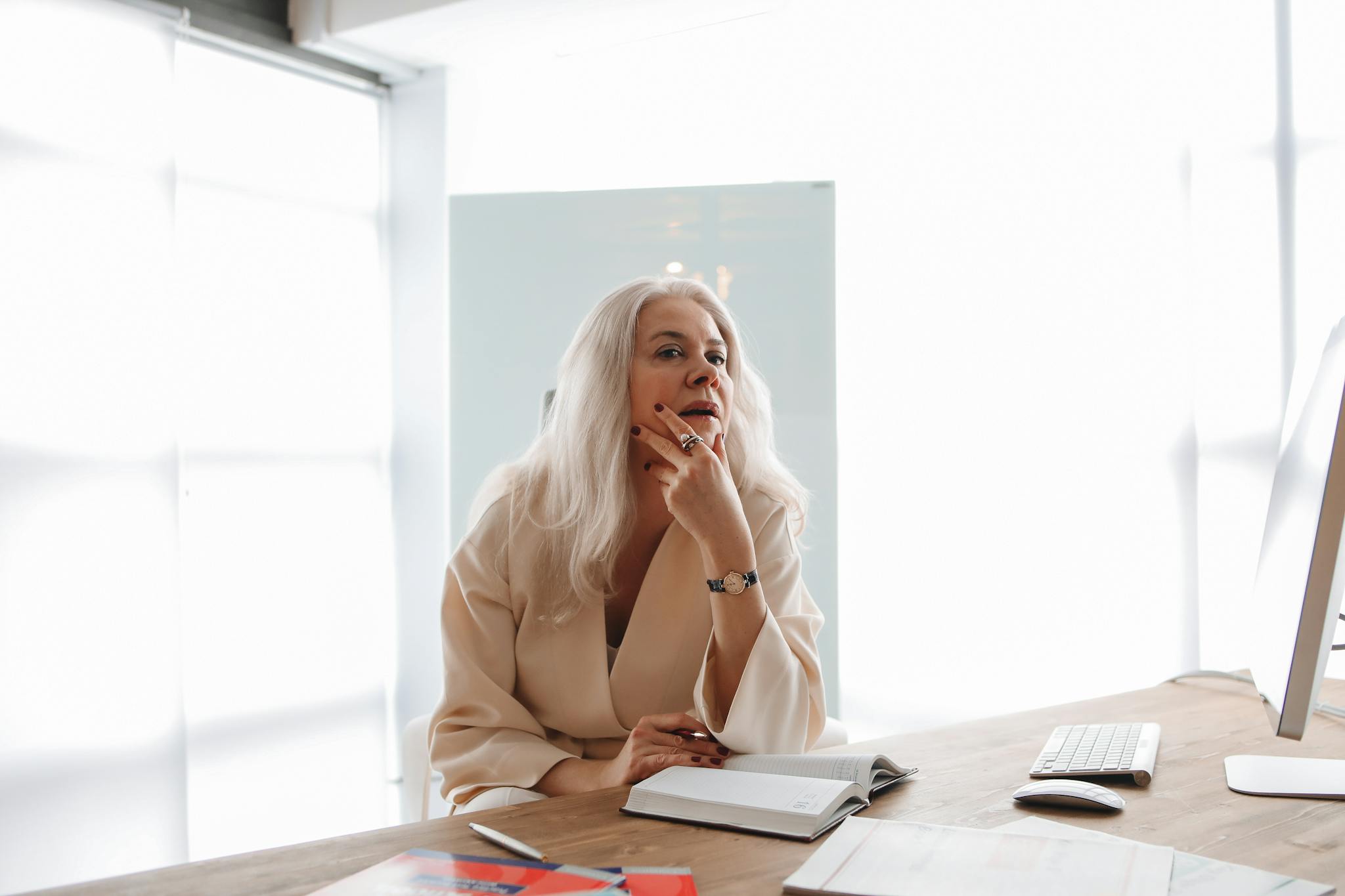 Senior woman thinking at a wooden desk while working on a computer with open books.